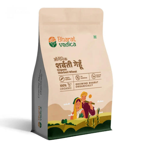 A beige and white packet of Bharat Vedica brand Organic Sharbati Wheat, with text in both Hindi and English. The packet features the brand logo at the top and an illustration of a farmer and a woman in a field of wheat at the bottom. The text on the packet reads "ऑर्गेनिक शरबती गेहूँ Organic Sharbati Wheat" and "100% ORGANIC GROWING BHARAT ORGANICALLY".