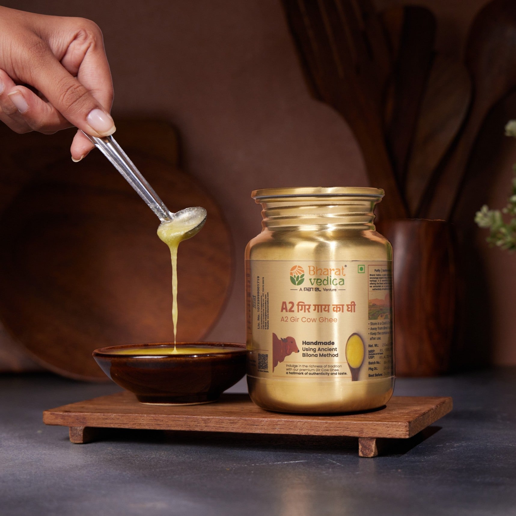 Stainless steel jar of 'Bharat Vedica' a2 gir cow ghee with a spoonful being poured into a bowl on a wooden stand.